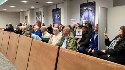 About two dozen people are sitting in the gallery of the council meeting, which is behind a wooden panel. They make up three rows of chairs between them. Some are holding signs. One reads: "No to city of sanctuary."
