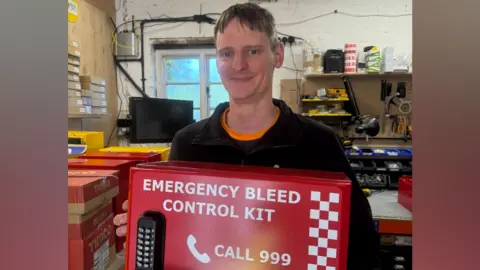 Annabel Amos/BBC Man in a black jumper and orange shirt poses with a bed emergency bleed control kit box.