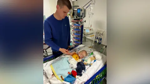 Family Alaric as a baby in a cot, surrounded by cuddly toys. He is in a hospital ward, and has tubes coming out of his nose. His father holds his hand and the room is full of medical monitoring equipment.