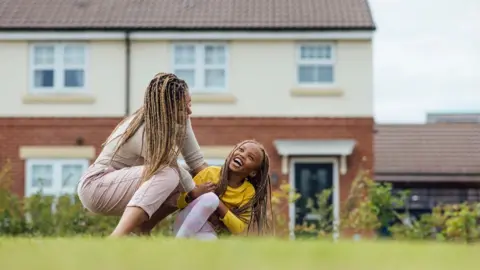 A mother plays with her daughter in a park in front of housing in the North East of England