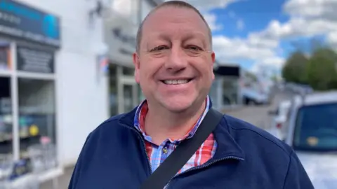 Darren Snelgar has very short brown hair and is wearing a blue fleece over a red and blue checked shirt. Behind him, out of focus, is a row of shops.