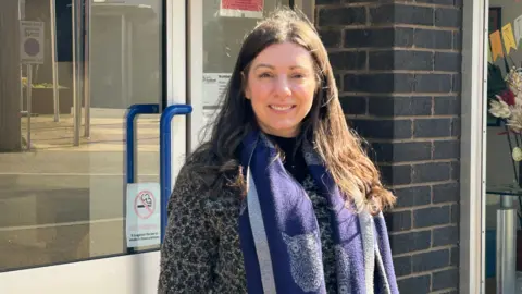 Gavin Kermack A woman with long dark hair standing in front of a door. She is wearing a purple scarf and is smiling at the camera as the sun shines on her face.