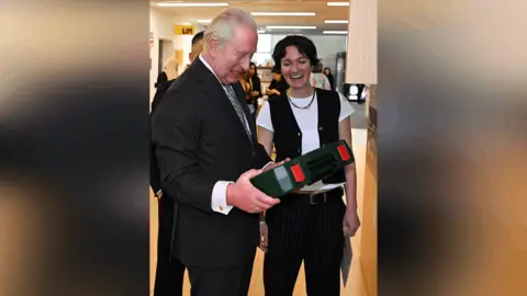 Getty Images King Charles holding a green case for an item that can be hired from the Library of Things. Next to him is a smiling person wearing a white t-shirt, black waistcoat and black trousers.