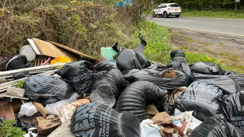 Bin baps, cardboard and other debris piled up on the side of a road. A white car drives in the background.
