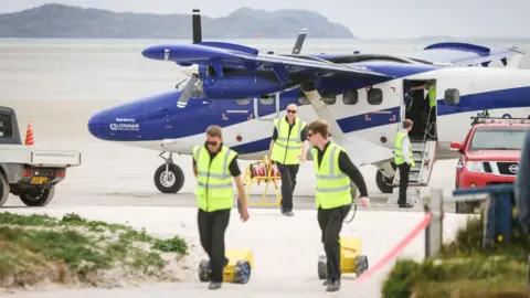 Three men in yellow high-vis vests wheel items in front of a blue and white airplane parked on a beach. In the distance, a black island can be made out. 