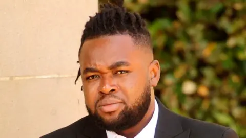 A man with a beard and dark hair, closely cropped at the sides, wearing a suit, pictured outside a court