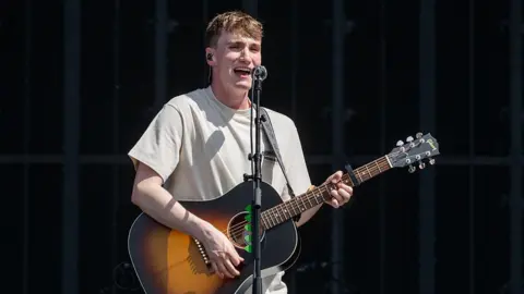 Getty Images Calum Bowie, a young singer with a sunburst Gibson acoustic guitar, stands at a microphone, singing. He is wearing a cream T-shirt and light trousers.