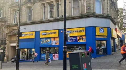 Cash Generator pawn shop with blue and yellow signage in a grand Grade II-listed building in Bradford city centre on the corner of Kirkgate and Bank Street, including pedestrians passing and a bin in the foreground.