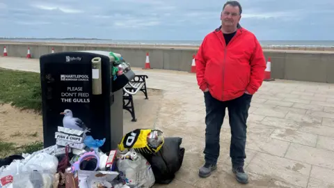 Paul Granger, a man wearing a red waterproof jacket and black trousers, with his hands in his pockets, is standing next to a large public bin overflowing with rubbish, and several bags of litter on the ground next to it.