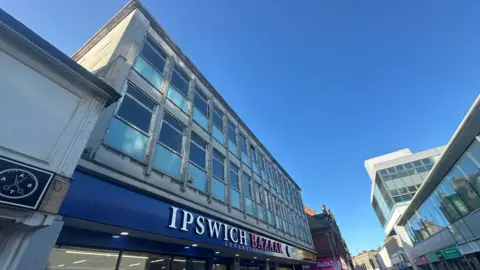 A view of the building where the 60 flats will be created in Ipswich. A large three storey grey building can be seen. Windows line the building. The Ipswich Bazaar Superstore can be seen on the bottom floor.