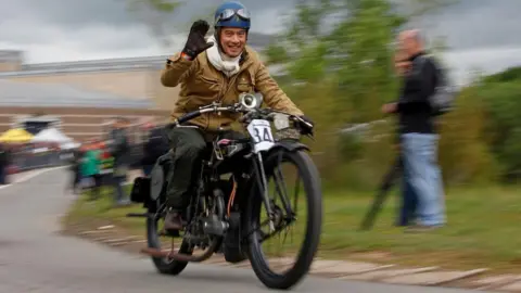 A man wearing a blue crash helmet with motorcycle goggles on it is riding a vintage motorcycle along a road. He is wearing a light brown bomber jacket and is smiling at the camera and waving a gloved hand.