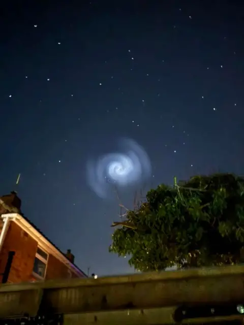 Emma Worship An image of a dark night sky with a bright white spiral in the centre. A brick house is on the left of the image and the sillhouette of a tree is on the right.