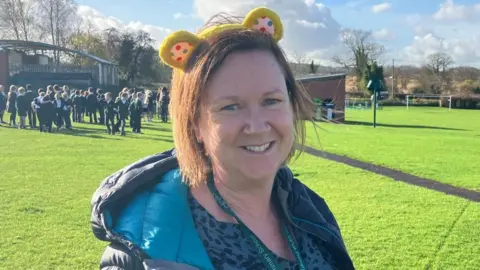 James Grant/BBC A woman with yellow bear ears smiles at the camera. Behind her in a school field, lots of children are grouped together talking. 