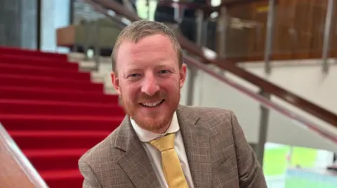 Newcastle City Council leader Colin Ferguson/ he is wearing a grey suit and yellow tie and has short brown hair and a short beard. He is standing on the stairwell inside Newcastle Civic Centre.