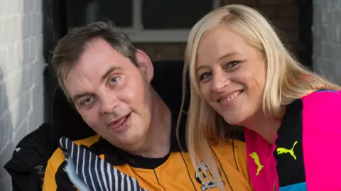 Cambridge United Simon Dobbin in a wheelchair wearing an orange and black Cambridge United strip. His wife, Nicole, is leaning in towards him and smiling. She is wearing a pink football top.