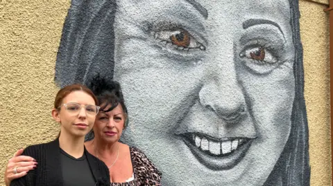 Deanna Ellis, who has light brown hair and glasses, and Sandra Bartlett, who wears a leopard print top and black shirt, stand in front of a mural by Jade Ellis