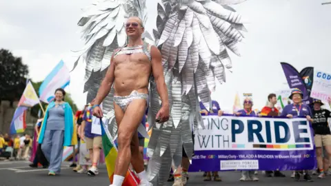 Matthew Horwood/Pride Cymru A man wearing silver thong has wings made of silver strips on his back as he walks in front of a crowd who are holding flags and banners