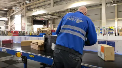 Guernsey Post staff member sorting parcels on a conveyer belt. The worker has a blue jacket on with the wording Guernsey Post on his back. He has a shaved head and is wearing black work trousers. He is facing away from the camera and we can see that he is in a large warehouse with white walls.