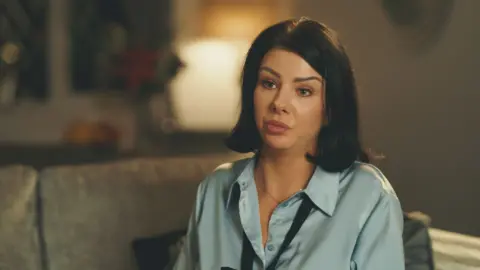 Laura Shearer sitting on a couch in a room. She has shoulder-length brown hair ansd is wearing a green blouse.