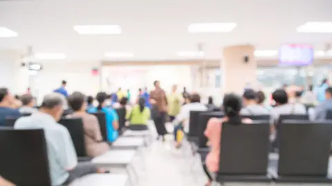 Getty Images A blurred image of a busy hospital waiting room. There are many people sitting on chairs. A TV can be seen in the distance. 