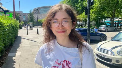 Sam Read/BBC Izzy stands wearing a white T-shirt with red writing on saying "Little Italy" and a large pair of pale red glasses. She has long brown wavy hair and smiles at the camera. In the background is a shopping centre and car park with trees and bushes.