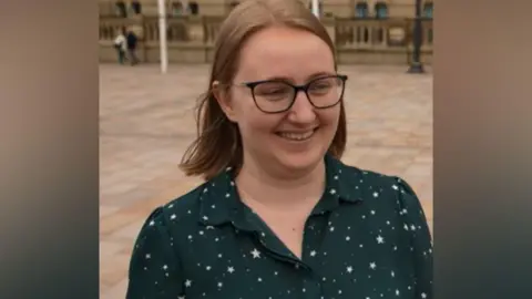 A woman with shoulder-length brown hair is pictured smiling while wearing thin framed black glasses. She is also wearing a dark green blouse with white stars dotted across it.