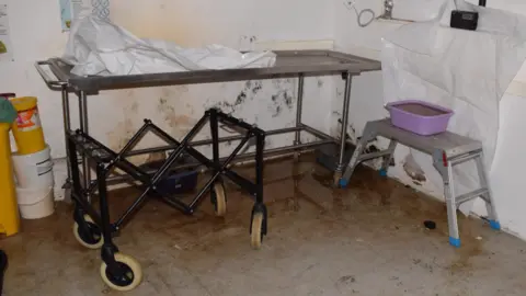 CPS A trolley in a mortuary. There is mould on the walls and the floor appears to be shiny with fluid.