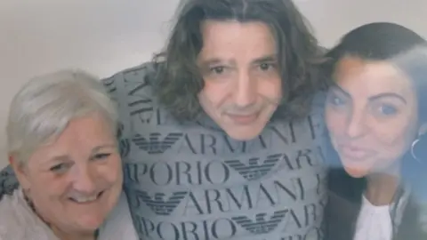 A middle-aged man with long dark hair stands with has arms around a younger and an older woman. 