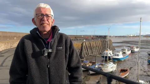 Allan is just off centre in the frame, and to the right of the photograph is the harbour with a few boats moored at low tide. Behind him is the harbour wall. Allan has white short hair and a pair of glasses. He's wearing a black fleece jacket.