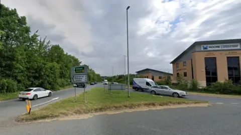 Single carriageway road seen from roundabout.  There are road signs to Oakham and other places. There are cars and vans on the road. An industrial unit is visible to the right.