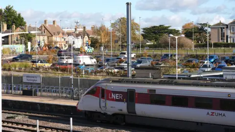 জো গিডেন্স/পিএ রেলওয়ে স্টেশন প্ল্যাটফর্মে একটি LNER ব্র্যান্ডের ট্রেন। প্ল্যাটফর্মে একটি চিহ্ন বলে "হান্টিংডনে স্বাগতম"প্ল্যাটফর্মের ওপারে স্টেশন কার পার্কিং এবং তার বাইরে বিল্ডিং।