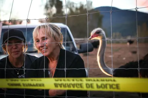 Aaron Hemens/The Canadian Press via AP Karen Espersen, her daughter, Katie Pasitne, and an ostrich stand behind a chain-linked fence cordoned off with yellow police tape