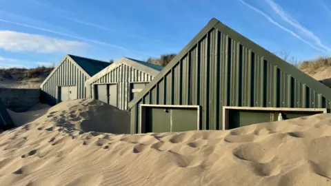 Rows of sand covered beach huts following storm, almost the same height as the doors.