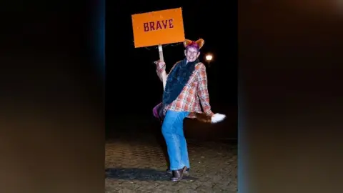 Jayne Jackson Photography A woman wearing an orange checked shirt, blue jeans, a pair of fox ears and a fox tail. She's striking a pose, smiling, and holding up a sign saying "brave".