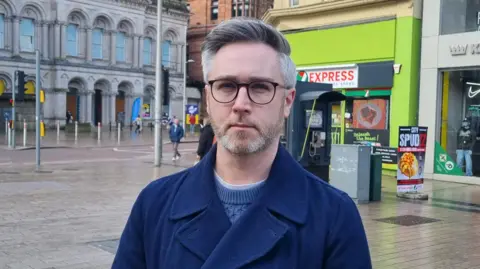 Man wearing glasses, a navy coast and light blue knitted jumper. He is standing in front of a telephone box and a fluorescent green convenience shop. 