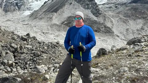 Simon Clark Simon Clark standing on rocks. He is wearing a blue shirt and a grey hat. He is leaning on walking sticks.