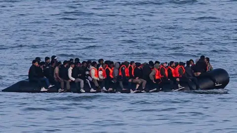 An overloaded inflatable boat sails through waves on a grey day. 