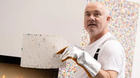 Getty Images Damien Hirst holds up a piece of art before burning it in a nearby fire. He is wearing a white t shirt, black braces, and a silver heat-proof glove.