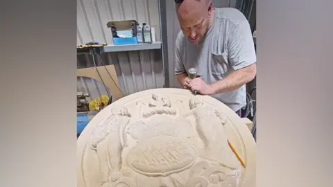 A bald man in a grey t-shirt is chipping away at a large white stone crest with masonry tools.