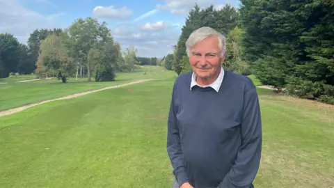 LDRS An elderly man stood on a golf course smiling into the camera. He is wearing a shirt underneath a navy blue jumper.