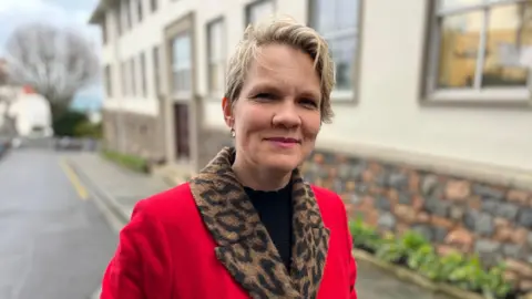 A woman with short blonde hair looking at the camera and smiling. She is wearing a red coat with a leopard-print collar.
