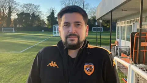 BBC Aaron has brown eyes, short dark hair and same coloured beard and moustache. He is wearing a black t-shirt under a black sports jacket bearing the orange Hull City logo on the right-hand side of the chest. 