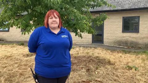 BBC/Phil Bodmer Emily standing outdoors in front of a leafy tree and a light-coloured stone or brick building. She is positioned slightly left of centre in the frame, facing the camera with both hands held behind her back.
She is wearing a bright blue polo shirt and dark trousers. A handheld radio is clipped to the waistband on the left side. The polo shirt has visible white text and a logo on the chest reading "RSPCA Sheffield". Her hair is short and reddish-orange in colour.