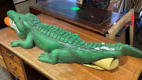 Alex Pope/BBC A crocodile prop laying on a wooden desk. It has a yellow pole going through it. Another table is behind it. 