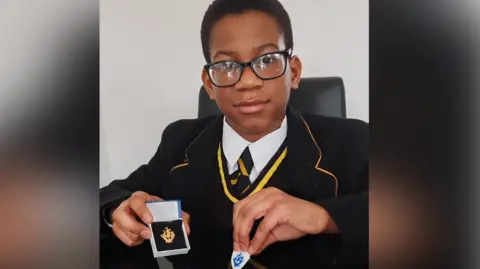 Anim Joyce A young boy in blame-rimmed glasses is wearing a black and yellow school uniform, sat at a desk holding two blue peter badges