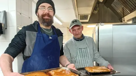 Other Two men in aprons hold up trays of pies