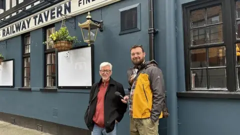 Two men stand in front of a blue-painted pub with a black and white sign that reads The Railway Tavern. The man on the left has short white hair and is wearing a black jacket over an orange top and blue jeans and black-framed glasses. Besides him stands a taller man with short dark hair and a beard, wearing a grey-sleeved yellow jacket and khaki coloured trousers. Both men smile towards the camera.