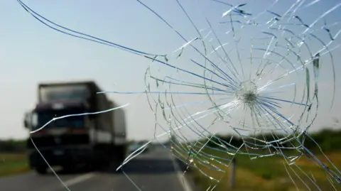 A large crack in a car windscreen. It is driving on a road and there is a lorry driving in the other direction.