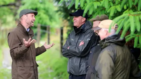PA Prince William smiling as he talks to three men who are wearing waterproofs and hats and sheltering from the rain under trees