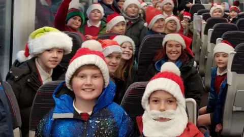 Children are crammed onto the train carriage. They are wearing Santa hats, scarfs and coats. One boy is wearing a false white beard. They are all looking at the camera